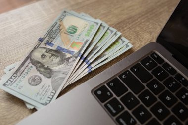 Tahta bir yüzeye dizüstü bilgisayarın yanına yerleştirilmiş yüz dolarlık banknotlar. Çevrimiçi kazanç, dijital finans ve uzaktan çalışma kavramı. Kaliteli fotoğraf. Yüksek kalite fotoğraf