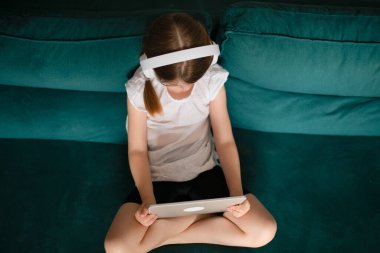 Kanepede elinde tabletle oturan küçük bir kız. Üst Manzara. İnternette çizgi film izleyen kız öğrenci..