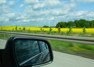 Otobandaki hareketli bir arabadan canlı sarı bir tarla, uzakta rüzgar türbinleri ve bulutlu bir gökyüzü görünüyor. Seyahat kavramı, yenilenebilir enerji, kırsal arazi ve çevre dostu taşıma
