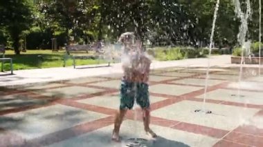 Güneşli bir yaz gününde halka açık bir parkta fıskiyeyle oynayan mutlu küçük çocuk. Su püskürtmelerinin keyfini çıkaran ve ıslanırken gülen bir çocuk. Açık şehir eğlencesi ve çocukluk sevinci.