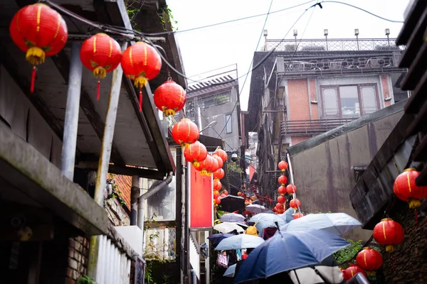 Tourists walk up the stairs with umbrellas as it rains, with red lanterns lining the houses along the way.