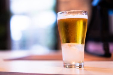 Soğuk altın biranın yakın planında, tahta bir masanın üzerinde temiz bir bardakta, bokeh light arka planda..