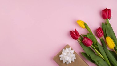 Beyaz fiyonklu bir hediye kutusu ve pastel pembe arka planda düzenlenmiş renkli laleler..