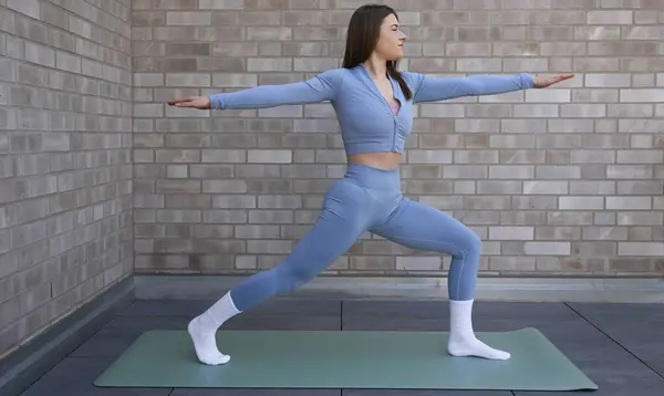 Genç bir kadın, çatıdaki terasta serilmiş yoga minderinde savaşçı pozu veriyor. Uyumlu mavi bir kıyafet giyiyor, parlak güneş ışığında yalın ayak, arka planda bir şehir silüeti var..