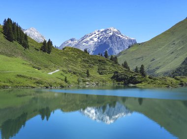 Parlak mavi bir gökyüzünün altında kristal berraklığında su ve karlı dağları olan manzaralı alp gölü.