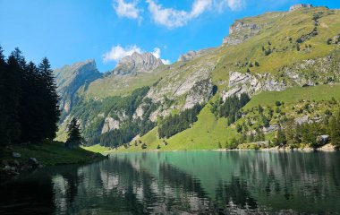 Güneşli bir günde, kayalık dağları ve yeşil yamaçları yansıtan kristal berraklığında suyu olan huzurlu dağlık bir göl.