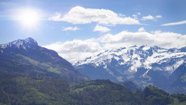 Karlı dağ zirveleri ve ilkbaharda yemyeşil vadileri olan panoramik alp manzarası. Parlak bulutlar ve mavi gökyüzü huzurlu doğa sahnesini tamamlıyor.