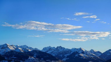 Karla kaplı dağ tepelerinin panoramik görüntüsü açık mavi gökyüzünün altında dağınık bulutlarla. Alpler 'in huzurlu kış manzarası, doğa ve seyahat temaları için mükemmel.