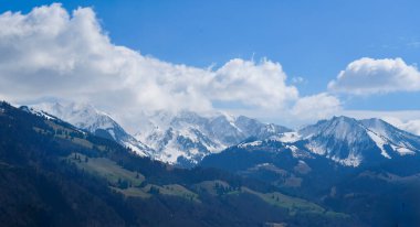 Karla kaplı dağ sırasının altında parlak mavi bir gökyüzü ve kabarık bulutlar. Yeşil vadileri ve görkemli zirveleri olan bahar dağları doğa ve seyahat temaları için mükemmel.