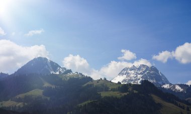 Açık mavi gökyüzünün altında karla kaplı iki dağ zirvesi olan parlak dağlık bir arazi. Yeşil tepeli bahar manzarası, orman yamaçları, ve tüylü beyaz bulutlar