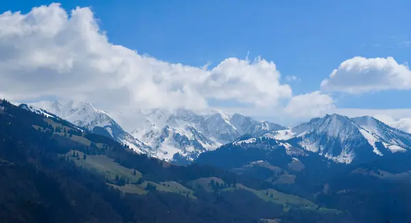 Karla kaplı dağ sırasının altında parlak mavi bir gökyüzü ve kabarık bulutlar. Yeşil vadileri ve görkemli zirveleri olan bahar dağları doğa ve seyahat temaları için mükemmel.