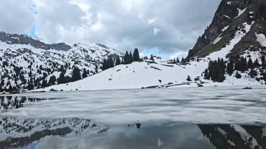 Bahar aylarında İsviçre Alplerindeki Seebergsee 'nin panoramik manzarası, karla kaplı yamaçlar ve dramatik bulutlu bir gökyüzü altında kısmen donmuş bir göl.