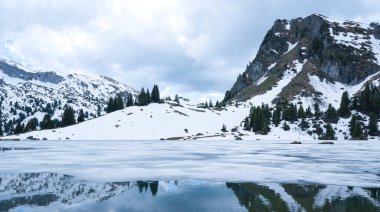 Seebergsee 'nin panoramik manzarası kısmen donmuş suyla kaplı, karla kaplı alp tepeleri ve bulutlu bir gökyüzünün altındaki kozalaklı ormanla çevrili.