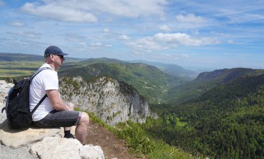 İsviçre Jura Dağları 'ndaki bir uçurumda dinlenen erkek yürüyüşçü, doğada yaz yürüyüşü sırasında panoramik dağ manzarasının tadını çıkarıyor.