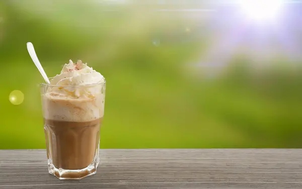 Krem şanti ve karamel ile buzlu kahve, ahşap bir yüzey üzerinde berrak bir bardak içinde sunulur, yumuşak yeşil arka plan ve sıcak güneş ışığı ile çevrili