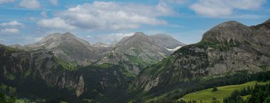 Çarpıcı dağ manzarası, yuvarlanan tepeler, kayalık zirveler ve parlak bir gökyüzü, doğal çevrenin güzelliğini vurgulayan barışçıl bir atmosfer yaratıyor.