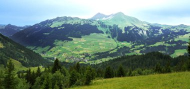 Yükselen tepeleri ve uzak dağlarıyla yemyeşil bir vadi, huzurlu bir doğal manzara yaratıyor, manzaranın ahengi ve açık hava sükunetini vurguluyor.