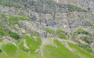 Kayalık dağ yamacında yemyeşil çimenler ve çarpıcı doğal manzarayı ve dağlık alanların eşsiz oluşumlarını vurgulayan ayrıntılı jeolojik dokular bulunur.