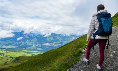 Kabarık ceketli ve sırt çantalı bir kadın dağ yolunda duruyor, göz kamaştırıcı yeşil tepelere ve uzak tepelere bakıyor, macera ve keşfin özünü yakalıyor.