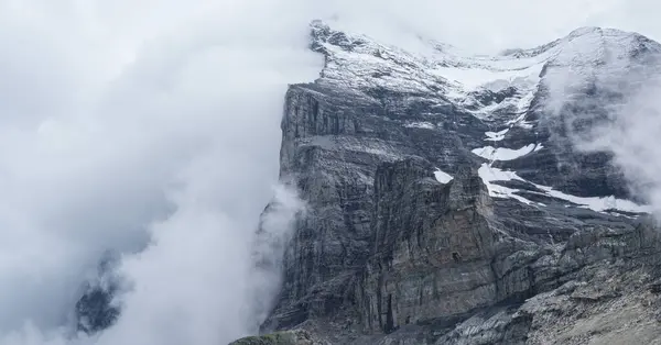 Bulutlarla kaplı dağ zirvesi engebeli uçurumları ve kar parçalarını ortaya çıkarıyor nefes kesen doğal ortamda sakin ve dramatik bir atmosfer yaratıyor.