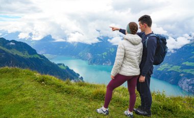 Çift İsviçre Alpleri 'ndeki çimenli tepenin üzerinde durur, aşağıdaki muhteşem dağ manzarasına ve sakin göle hayran kalır, bulutlar havada süzülür, sakin bir açık hava ortamı yaratır.