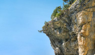 Parlak mavi gökyüzüne karşı yeşil ağaçlarla kaplı görkemli kayalık kayalıklar doğanın güzelliğini ve dışarıdaki manzaranın ihtişamını vurguluyor.