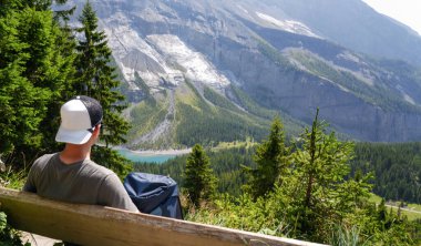 Bir bankta huzurlu bir anın tadını çıkaran, canlı ağaçlar ve sakin bir göl ile çarpıcı dağ manzarasına bakan, açık hava keşiflerinin güzelliğini somutlaştıran bir insan.