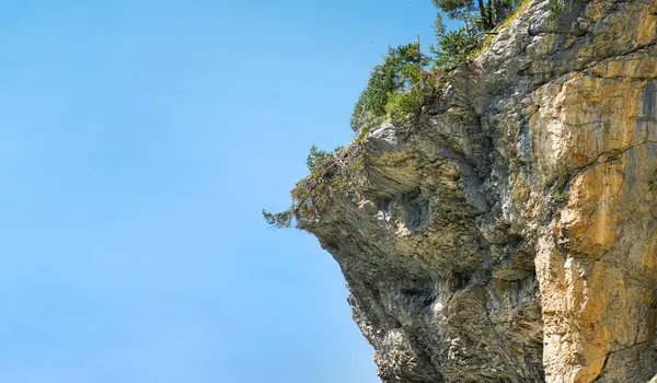 Parlak mavi gökyüzüne karşı yeşil ağaçlarla kaplı görkemli kayalık kayalıklar doğanın güzelliğini ve dışarıdaki manzaranın ihtişamını vurguluyor.