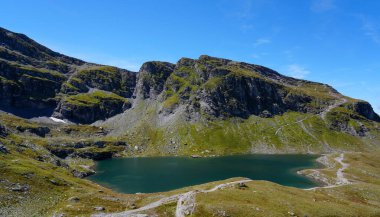 Schwarzsee 'nin manzarası, İsviçre' de bir dağ gölü, açık mavi gökyüzünün altında yeşil tepeler ve kayalık kayalıklarla çevrili.