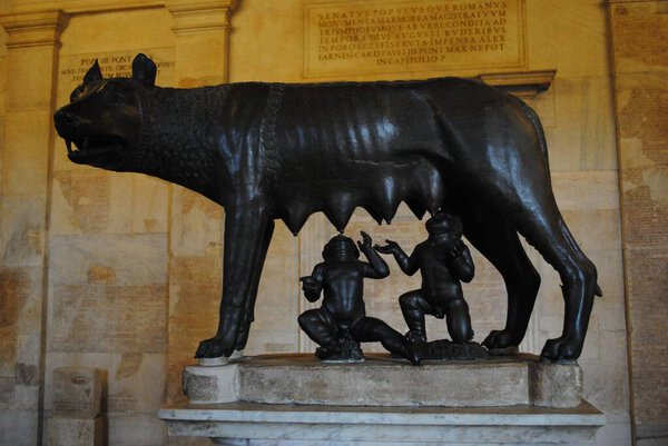 Bronze sculpture of the Capitoline Wolf suckling Romulus and Remus, displayed in a museum with a historical Latin inscription on the wall in the background.