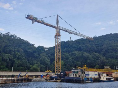 Yüksek bir endüstriyel vinç, yemyeşil ormanlarla çevrili bir nehir kenarı şantiyesine hükmediyor. Tekneler ve mavnalar, doğa ve endüstrinin ahenkli bir karışımı olarak berrak mavi bir gökyüzünün altındaki nehir kıyısına dizilirler..
