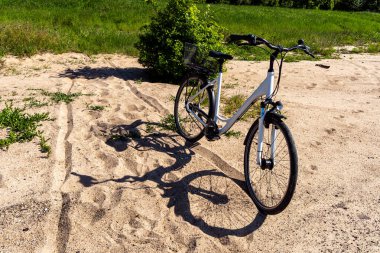 Kumlu bir patikada duran sepetli beyaz bisiklet, parlak güneş ışığında bir gölge bırakıyor, arka planda yeşil çayırlar var..