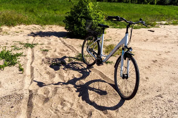 Kumlu bir patikada duran sepetli beyaz bisiklet, parlak güneş ışığında bir gölge bırakıyor, arka planda yeşil çayırlar var..