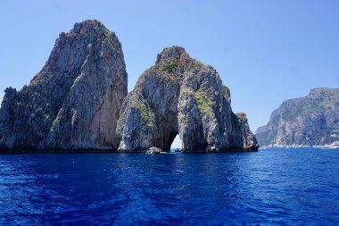 Capri, İtalya açıklarındaki ikonik Faraglioni kaya oluşumlarının çarpıcı bir görüntüsü. Bu yüksek kireçtaşı kayalıkları, engebeli yüzeylerini süsleyen canlı yeşillikleriyle derin mavi Akdeniz 'den çarpıcı bir şekilde yükselir..