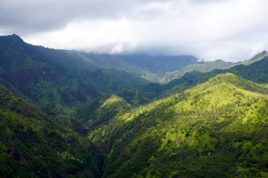 Bulutlu bir gökyüzünde gün ışığıyla aydınlanan yemyeşil dağların nefes kesici manzarası. Yuvarlanan tepeler ve derin vadiler doğanın güzelliğini ve zenginliğini gösteren dinamik ve canlı bir manzara yaratıyor..                               