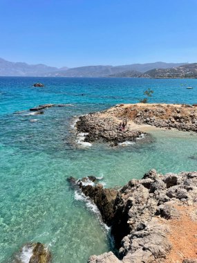 Turkuaz sular, Yunanistan 'ın Girit kentindeki kayalık bir sahilde, parlak bir güneş ve berrak mavi bir gökyüzü altında, huzurlu ve resimli bir sahil cenneti yaratarak nazikçe tur atıyor.