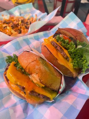 İki yağlı duble çizburger, marul domatesli ve soslu altın brioche çörekleri damalı mavi-beyaz kağıda oturup yanında patates kızartması ile leziz ve hoşgörülü bir yemek yaratıyor.