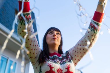 Young woman hanging christmas lights outdoor on a sunny day clipart