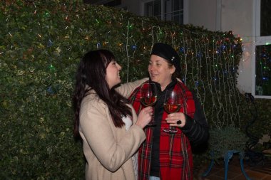 İki kadın gülümsüyor ve yılbaşı ışıklarının önünde şarap kadehleri tutuyorlar.