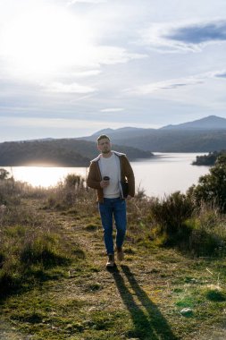 Genç adam gölde huzurlu bir yürüyüşün tadını çıkarıyor, elinde bir kahve ve defter, doğanın huzurlu güzelliğini kucaklıyor.