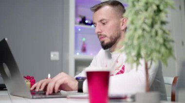 Gri bir dizüstü bilgisayarla evden telgraf çeken genç bir adam, Noel süsleriyle çevrili, daktilo ediyor ve gülümsüyor. Yüksek kalite 4k görüntü