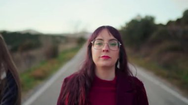 Uzun kumral saçlı ve gözlüklü bir kadın köy yolunda dikilirken yan tarafa bakıyor.