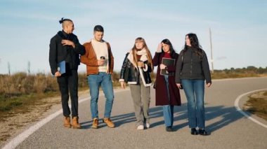 Dört üniversite öğrencisi asfalt yolda yürüyor, sohbet ediyor ve kahvelerini yudumluyorlar.