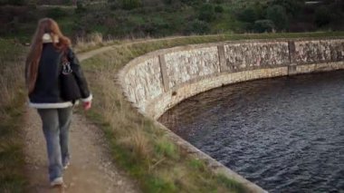 Uzun saçlı ve siyah ceketli bir kadın su deposunun yanında yürüyor.