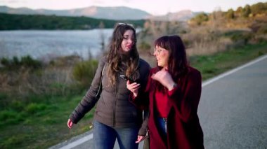 İki arkadaş göl kenarındaki manzaralı bir yolda yürüyorlar, sohbet ediyorlar ve manzarayı gösteriyorlar.