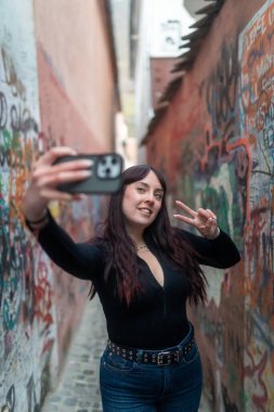 Turist grafiti dolu bir sokakta selfie çekiyor, barış işareti yapıyor ve kameraya gülümsüyor.