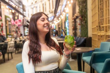 Elinde mojito kokteyli tutan genç bir kadın, Noel tatilinde dekore edilmiş bir açık hava barında bir masada oturuyor.