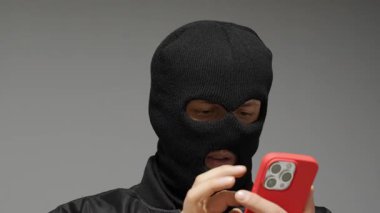 Gri arkaplandaki maskeli hacker akıllı telefonu tarıyor