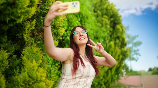 Mutlu genç bir kadın akıllı telefonuyla selfie çekiyor ve güneşli bir günde parkta barış işareti yapıyor.
