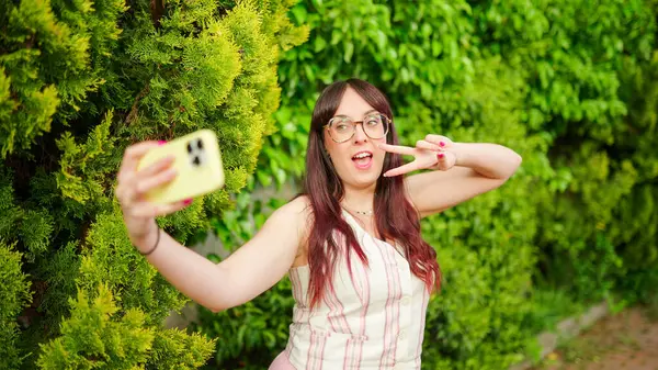 Neşeli genç bir kadın akıllı telefonuyla selfie çekiyor, parmaklarıyla zafer işareti yapıyor.